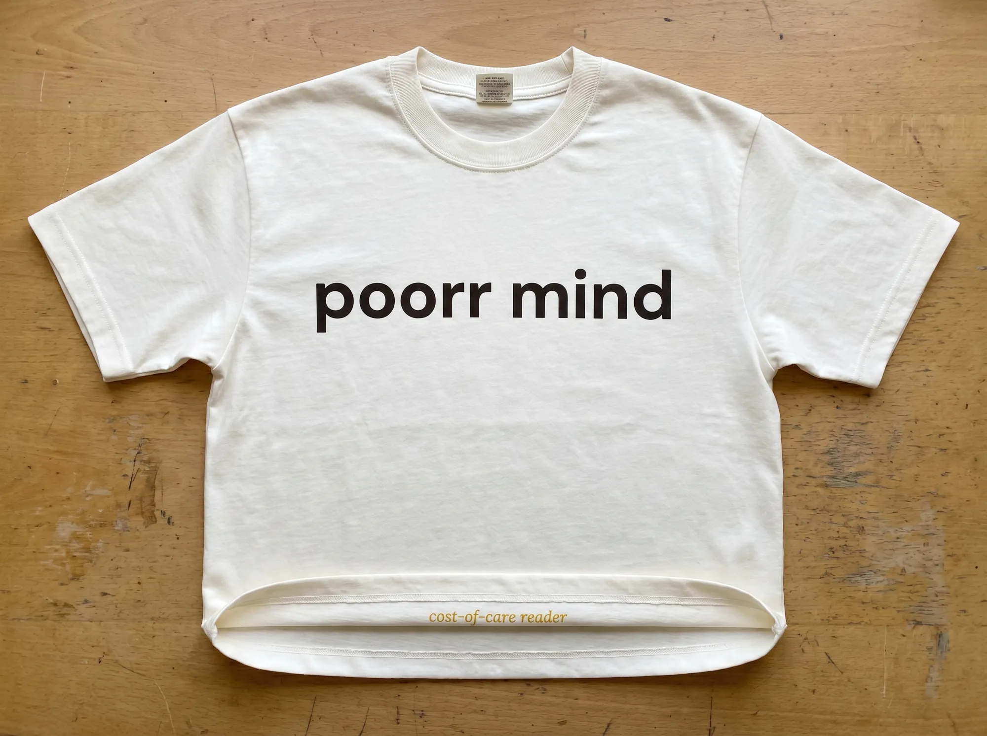 Poorr Mind Tee (Apparel) — A heavyweight cream cotton tee with the wordmark in warm charcoal and a small Lora italic line at the hem reading 'cost-of-care reader.' For Mind subscribers.