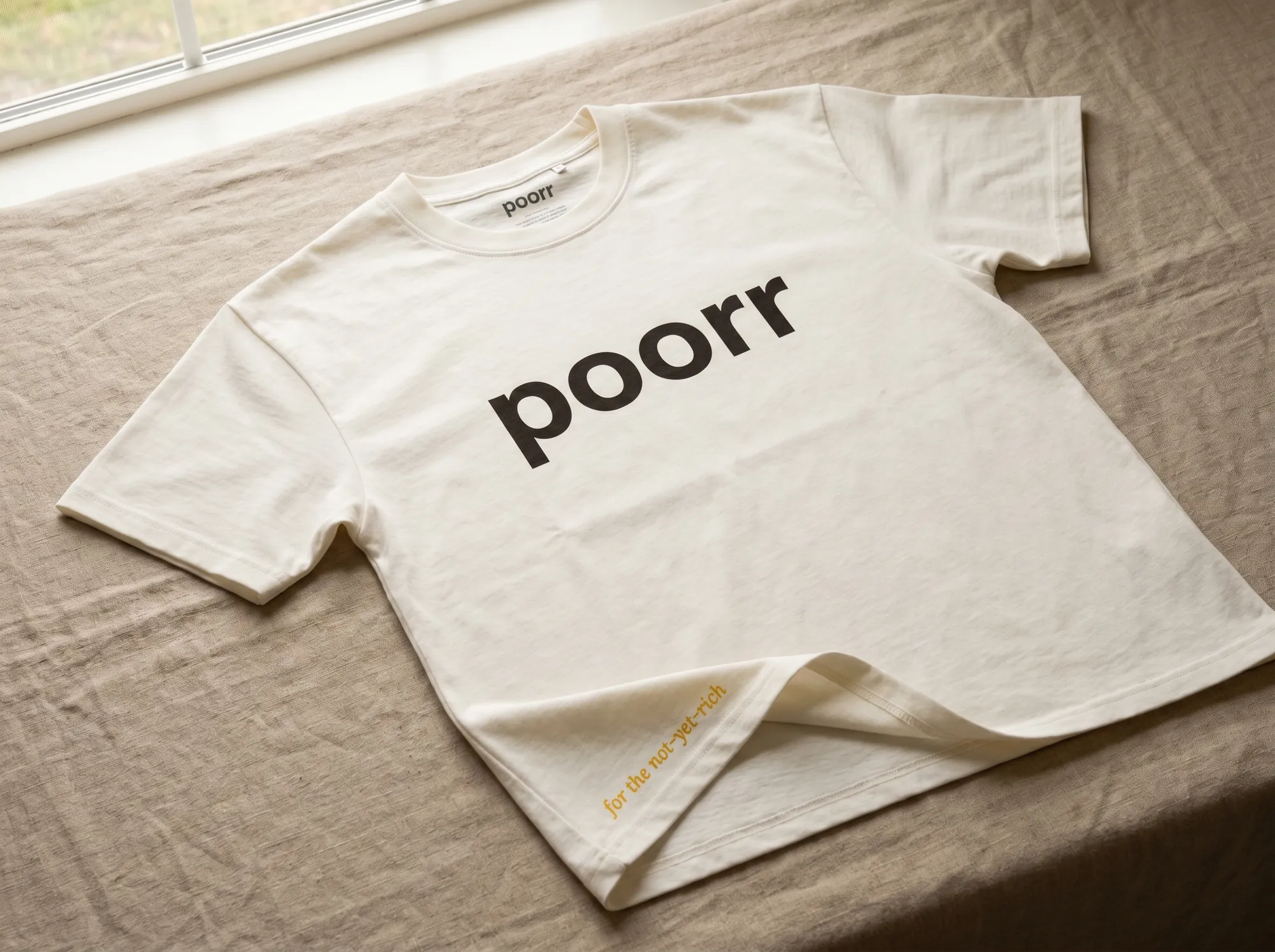 Poorr Tee (Apparel) — A heavyweight warm off-white cotton tee printed with the wordmark in warm charcoal and a small Lora italic line at the hem reading 'for the not-yet-rich.' Plain B2C apparel.